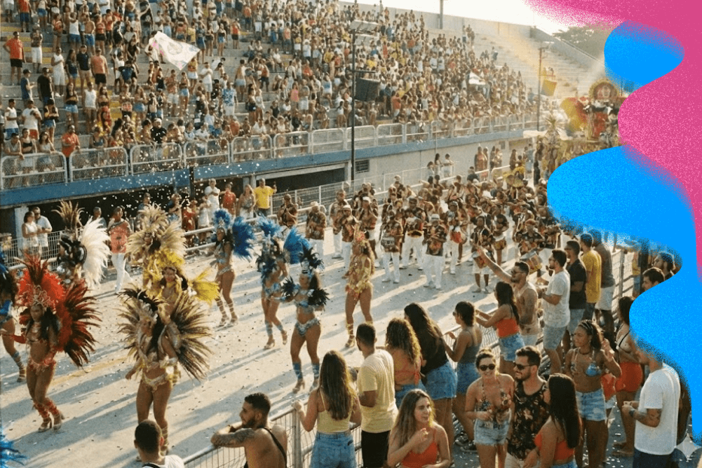 Desfile de escola de samba no Sambódromo com passistas em fantasias de plumas, bateria e público nas arquibancadas ao fundo.