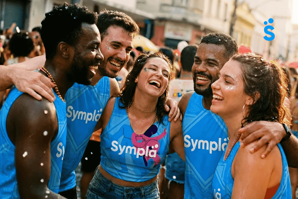 Grupo de amigos sorrindo e abraçados em um bloco de rua, usando abadás azuis da Sympla personalizados para o Carnaval.