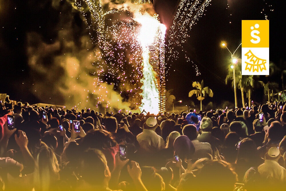 A imagem mostra uma grande produção de festa junina. O público observa uma grande fogueira à noite.