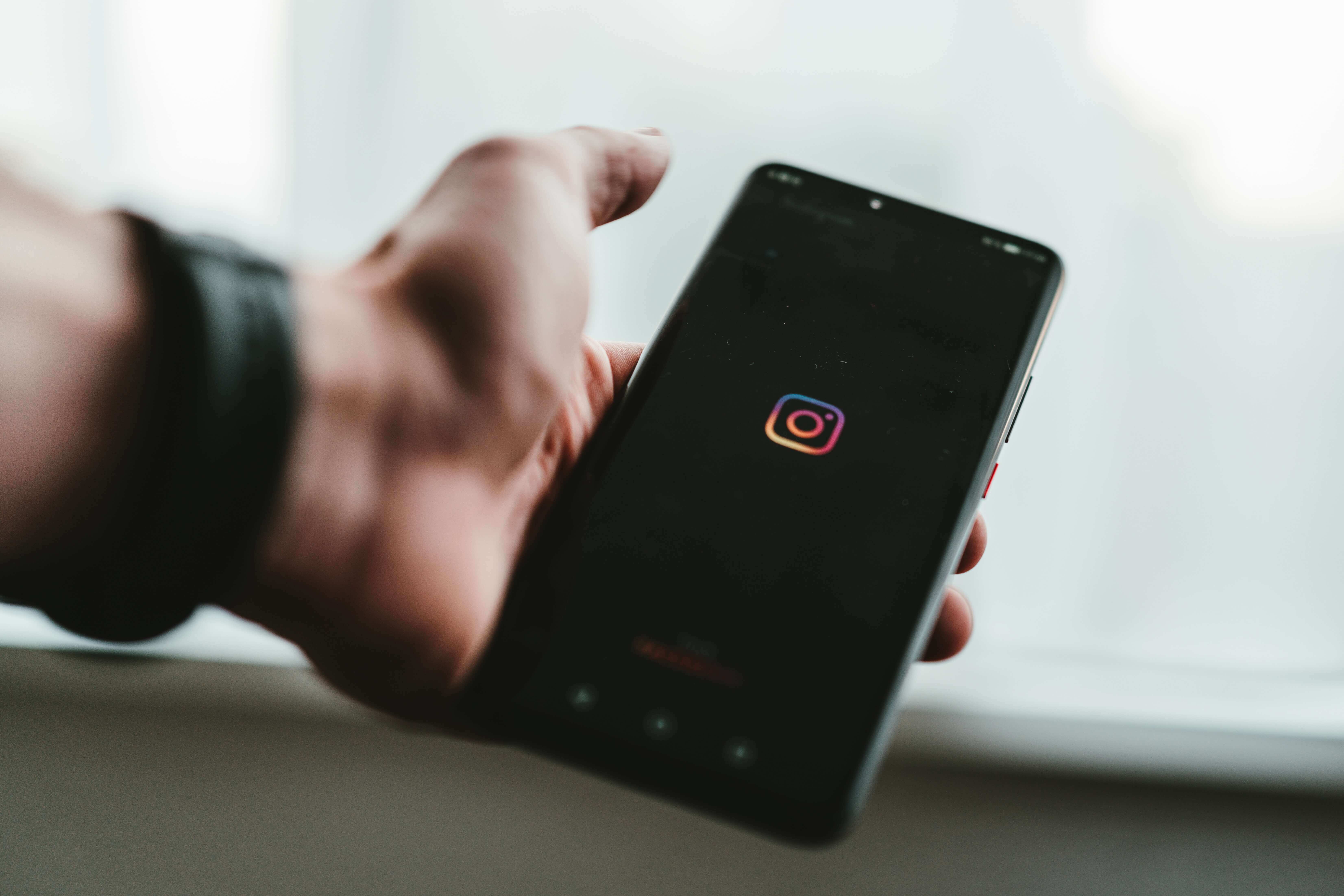 A imagem mostra uma mão segurando um telefone celular. Na tela, há um fundo preto e a logo do Instagram.