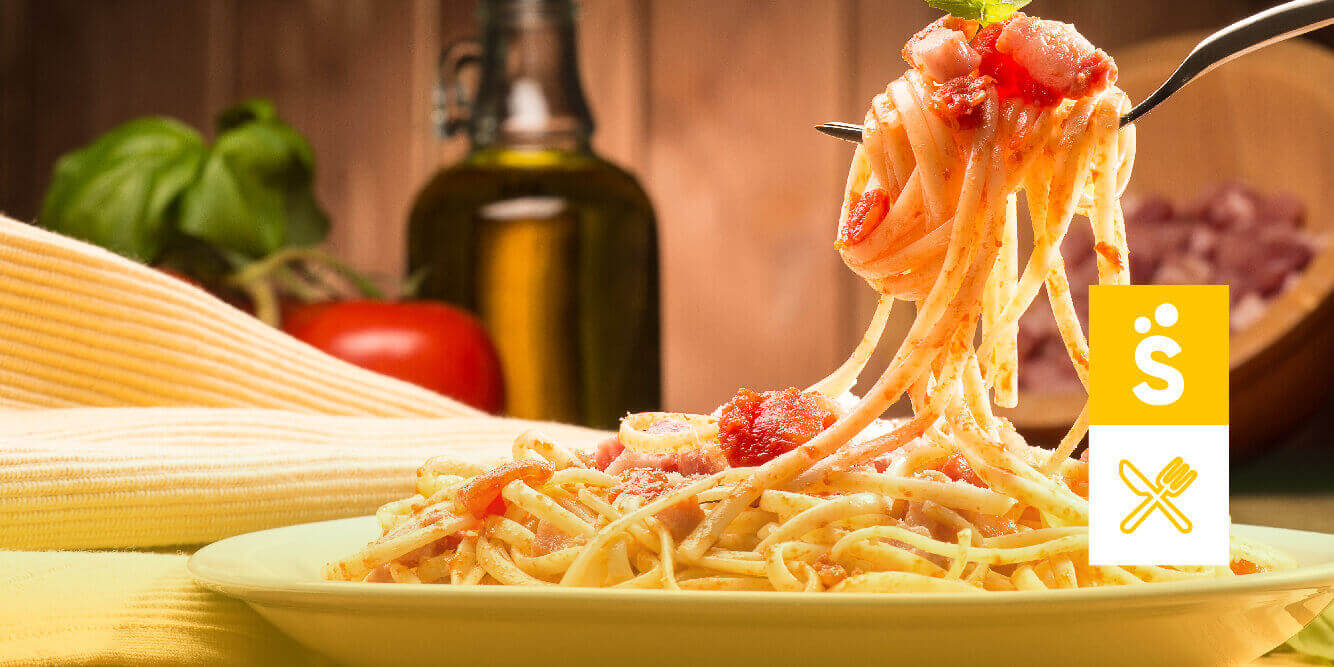 Imagem ampliada de um prato de macarrão com elementos ao fundo, como jarra de azeite e tomate