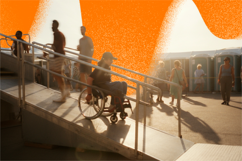 Pessoa usuária de cadeira de rodas desce uma rampa de acesso em um evento ao ar livre, com corrimão metálico. Outras pessoas circulam ao fundo, onde há banheiros químicos azuis visíveis. A cena parece ocorrer no entardecer, com luz quente, e há uma grande área/sobreposição gráfica laranja na parte superior da imagem.